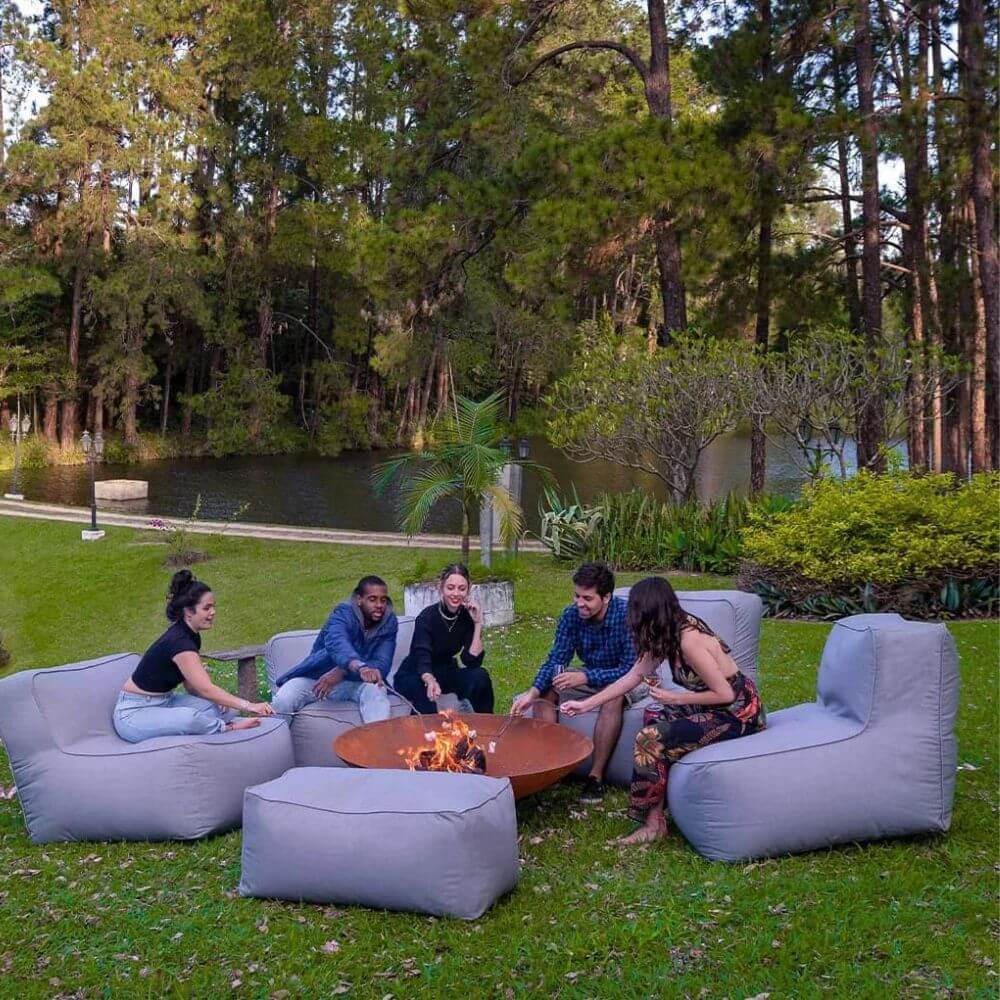 Grupo de amigos reunidos ao redor de uma fogueira em um espaço verde, com puffs confortáveis, criando um ambiente descontraído para o Réveillon.. Entenda tudo sobre como decorar sua festa de ano novo!
