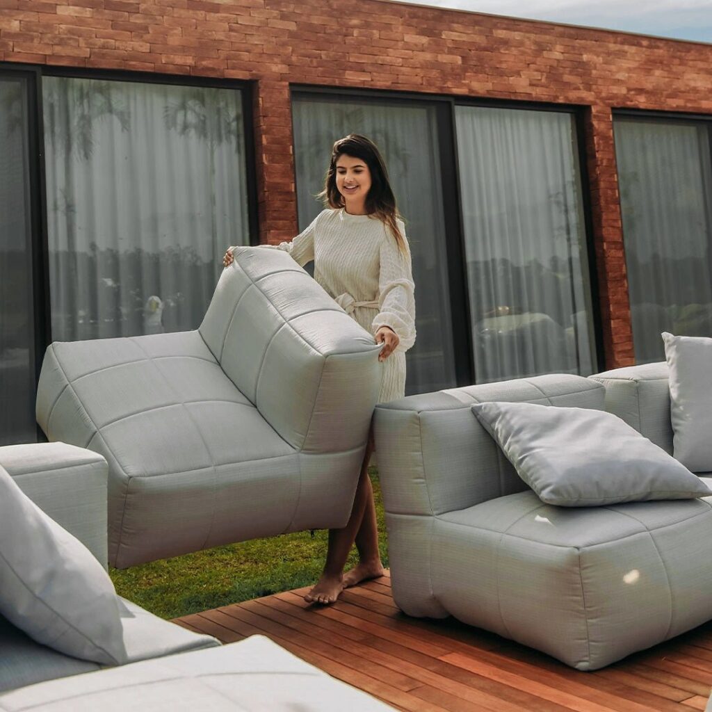 Mulher ajustando sofás modulares cinzas em um deck de madeira, mostrando versatilidade na decoração para jardim externo.